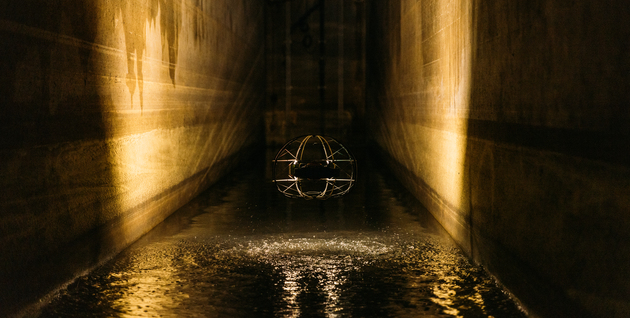 ASIO X drone with lights on hovers over water in in sewer, its propellers creating ripples on the water's surface.