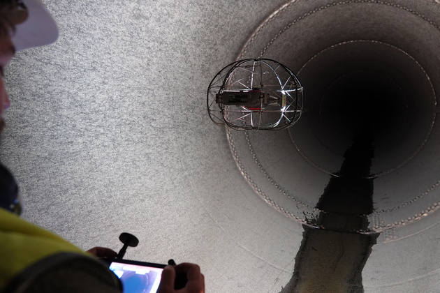 An ASIO X drone is flown inside a penstock, viewed over the shoulder of a pilot wearing a safety vest and operating the drone with a remote control.